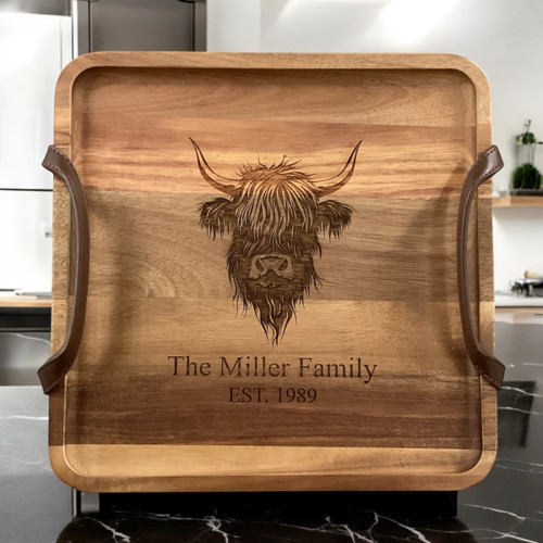 A square wooden serving tray featuring a dark brown laser-engraved illustration of a Highland cow head in the centre. The tray has a natural wood grain finish and is equipped with two dark brown leather handles attached to the sides.