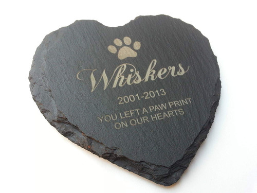 A heart-shaped black slate plaque with rough, uneven edges. The surface is engraved with a paw print icon, the name Whiskers, the dates 2001-2013, and the text YOU LEFT A PAW PRINT ON OUR HEARTS in a light-coloured, sans-serif font.