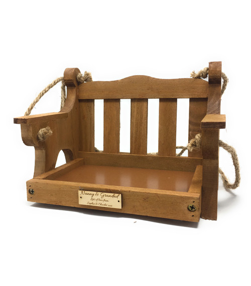 A wooden bird feeder in the shape of a swing, with a slatted back and a tray base. It is suspended by rope and has a small plaque with text. The feeder is made of light brown wood. Engraved message plaque on front creating a unique garden gift.
