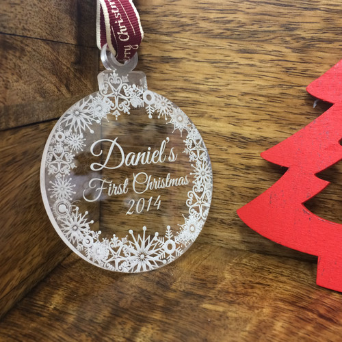 A clear 5mm acrylic Christmas bauble featuring an engraved snowflake border and the text Daniel's First Christmas 2014. The round decoration includes a red ribbon for hanging and is displayed with detailed circular insets.