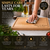A rectangular light brown wooden chopping board with a juice groove, engraved with the text Chris & Alison and 18th July 2026. The board is being wiped with a checkered cloth, featuring instructional text about care and maintenance.