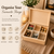 A light-coloured wooden storage box with a hinged lid and six removable compartments. The box contains tea bags, small gold-wrapped sweets, dried flowers, small photographs, and a string of white pearls. Text reads: Organise Your Favourite Things.