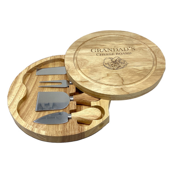 A round wooden cheese board with a swivel lid engraved with the text GRANDAD'S CHEESE BOARD and a graphic of cheese. The board opens to reveal a set of four stainless steel cheese servers with wooden handles.