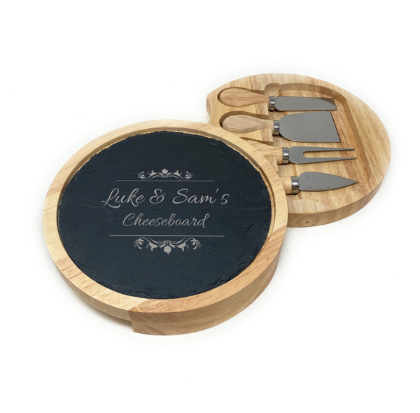 A round wooden cheese board with a removable black slate top. The slate is engraved with the text: You're the Best Daddy Ever, Love, Emily & Gracie, x 2026 x. The board features a hidden drawer for storing four stainless steel cheese tools.
