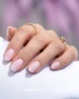 Elegant manicure featuring softly rounded almond-shaped nails in a delicate milky pink shade with a smooth, glossy finish. The image highlights a natural-looking soft pink manicure complemented by delicate gold rings, white fabric and a subtle lilac background, showcasing the refined nail appearance associated with Makear 603 Milky Pink Hybrid Gel Polish.