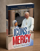 Three-quarter view of the Icons of Mercy paperback on a wooden surface, showing the front cover, spine, and page edges.