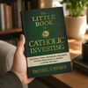 An adult holds The Little Book of Catholic Investing in one hand with the front cover facing the viewer.