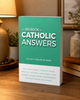 The Big Book of Catholic Answers, Volume 3: The Life of Faith, standing on a wooden tabletop beside a warmly lit reading lamp.