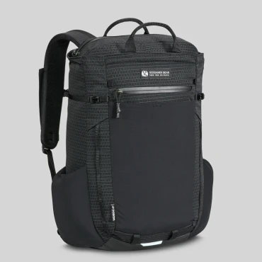 Looking at the corner of the backpack standing up against a white backdrop. The backpack is black and rectangular and is shown to have a front waterproof zip, 2 vertical daisy chains, and a large front mesh pouch on the bottom three quarters of the backpack. Also pictured are the large carry handles on top of the backpack.