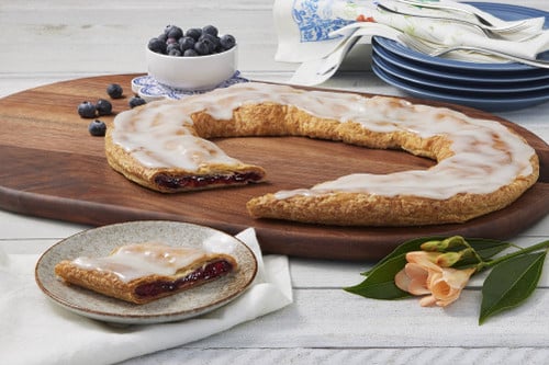 Blueberry Kringle sitting on a wooden cutting board with a piece cut out and some blueberries in a small white bowl near the kringle.Blueberry Kringle sitting on a wooden cutting board with a piece cut out and some blueberries in a small white bowl near the kringle.