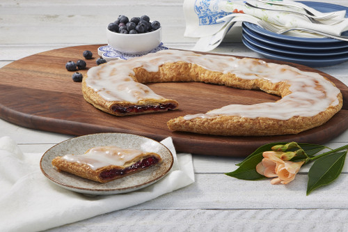Blueberry Kringle sitting on a wooden cutting board with a piece cut out and some blueberries in a small white bowl near the kringle.Blueberry Kringle sitting on a wooden cutting board with a piece cut out and some blueberries in a small white bowl near the kringle.