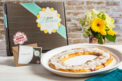 Brown box with turquoise stripe with "Just Because" font behind a kringle on a white plate next to a vase of yellow flowers and a brick wall background.