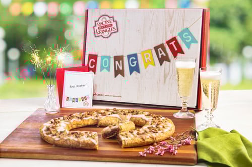 Congratulations Gift Box with a Kringle sitting on a wooden cutting board in front of the box with champagne and flowers near the kringle.