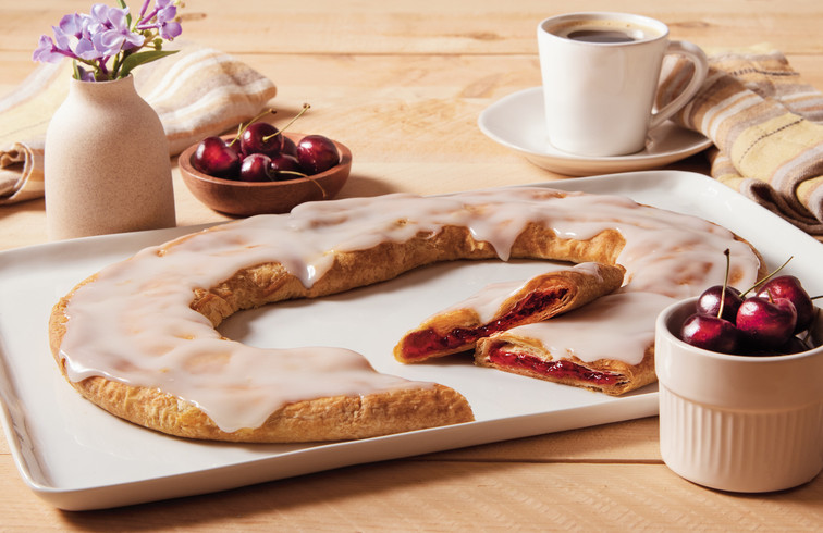 Cherry Kringle on a white plate on a wooden table with coffee and cherries next to the Kringle.