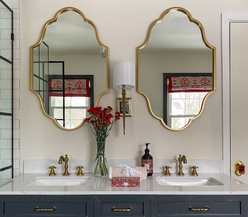 Clifton Wall Sconce by Crystorama styled in between two gold vanity mirrors above a marble top vanity with blue cabinet doors and gold hardware.