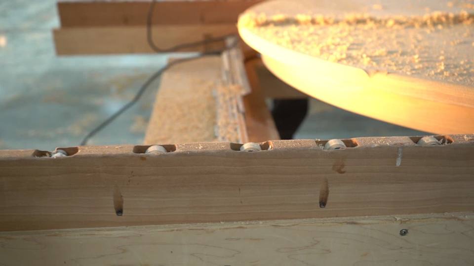 A handheld router is guided around the curved edge of a supported wooden tabletop. Shavings are visible on the surface at the beginning.