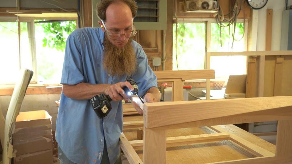 A worker uses a handheld driver near a corner of a wooden frame supported on a workbench, then shifts position.