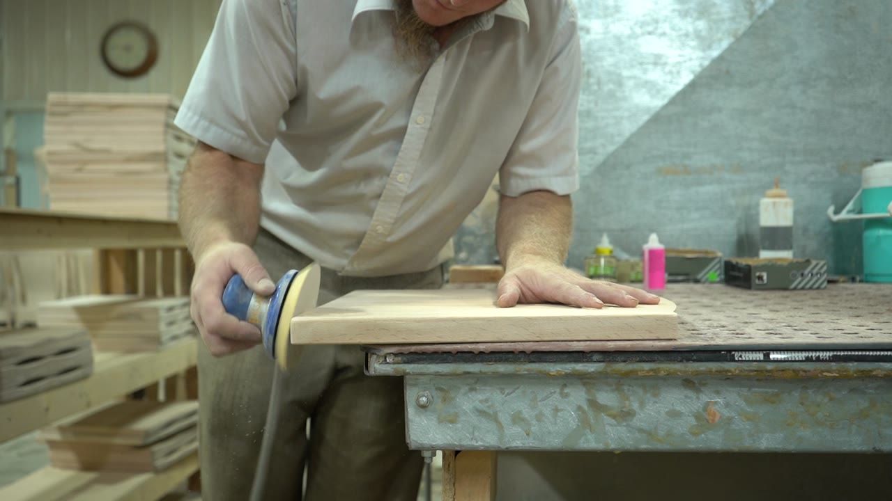 A worker steadies a shaped wooden chair seat while guiding a handheld sander along its edge