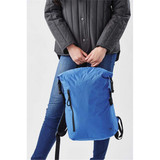 A blue roll-top backpack being held by a person wearing a grey jacket, with a logo visible on the bag.