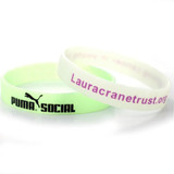 Two glow-in-the-dark silicone wristbands, one in pale green and the other white, featuring a logo on the green band.