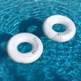 Two white inflatable swim rings floating in a blue pool, featuring the words "BRUNETTE" and "BLONDE."