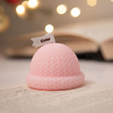 A pink hat-shaped candle with a textured, knitted design, placed on a book with blurred lights in the background.