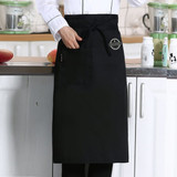 A black polyester canvas half apron with a pocket, featuring a logo, displayed against a kitchen backdrop.