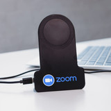 A black wireless charger with a circular charging pad and a logo, positioned next to a laptop.