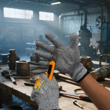 Cut resistant gloves in grey with a hand gripping a utility knife near various tools and materials in a workshop setting.