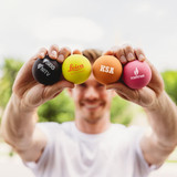 A person holds four colourful high bounce balls in various shades: black, yellow, orange, and pink, each with a logo.
