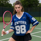 A young woman poses in a blue jersey featuring white text and a number, holding a pink tennis racket on a court.