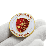 A gold and red enamel lapel pin featuring a shield design held on a white background.