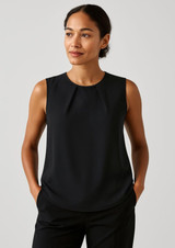 A sleeveless black blouse featuring pleat details, modelled by a woman with her hands in pockets.