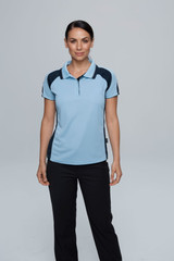 A woman stands wearing a light blue polo shirt with navy accents, paired with black pants. The shirt features a logo.