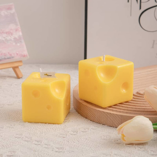 Square candles shaped like cheese, featuring a yellow colour with holes, displayed on a wooden plate with flowers nearby.