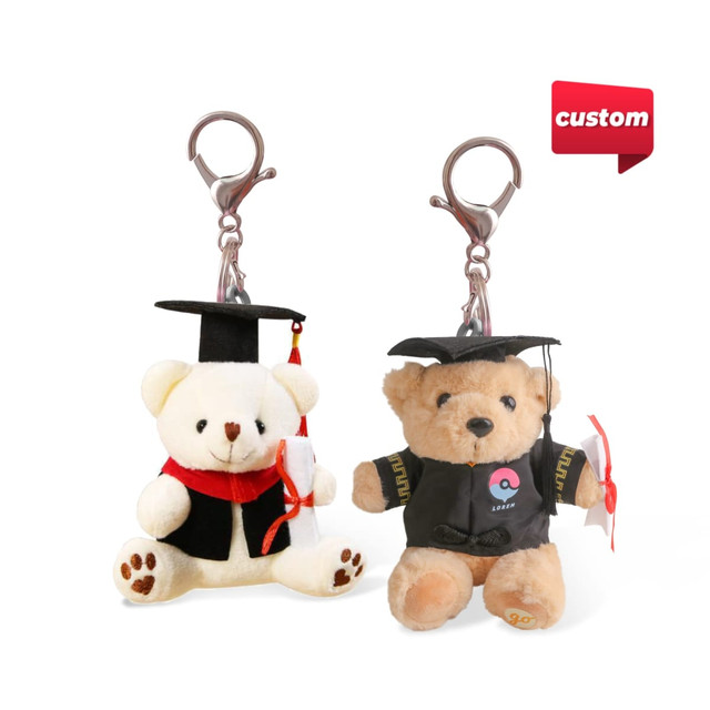 Two plush bear keyrings are dressed in graduation caps and gowns, featuring a tan bear and a cream bear, each with a clip.