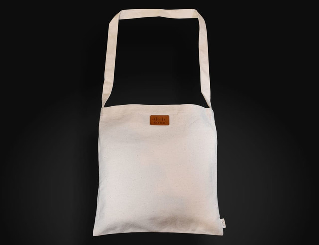 A light-coloured, long-handled shoulder bag with a logo on the front, featuring a simple, minimal design.