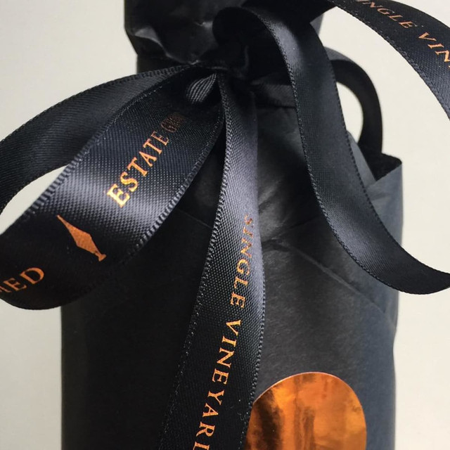 A bottle wrapped in black paper, topped with black satin ribbon featuring woven edges and a logo.