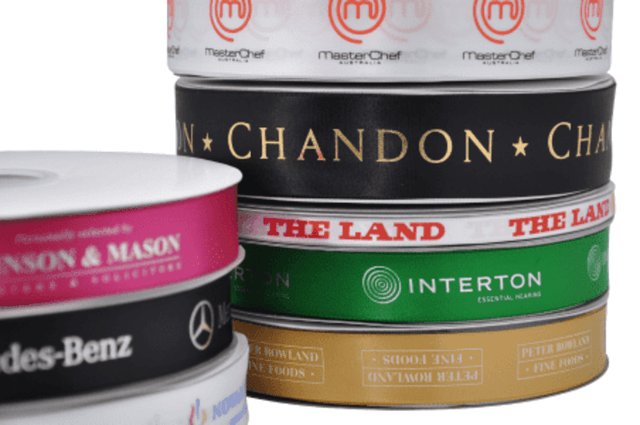 A stack of various custom printed double-sided satin ribbons in colours like black, pink, white, and green, with logos.