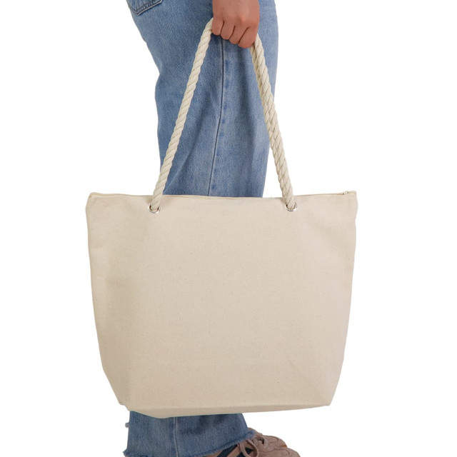 A cream canvas tote bag with rope handles, held by a person wearing denim jeans.