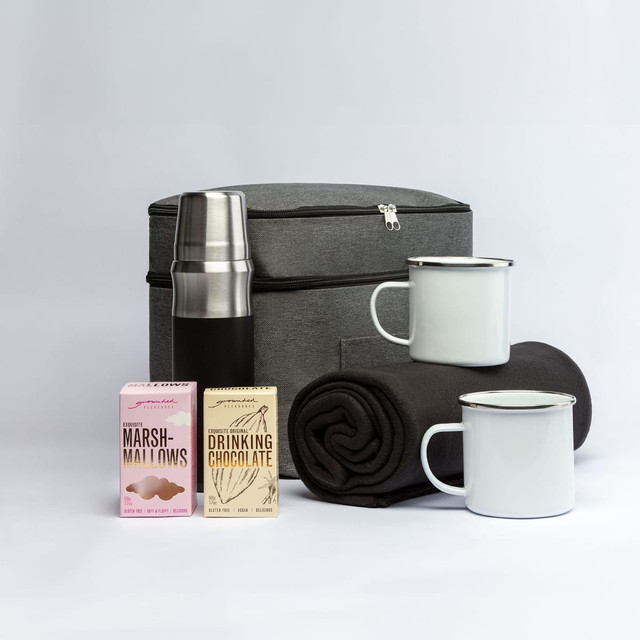 A grey cooler bag with a stainless steel drink bottle, two mugs, a blanket, and boxes of marshmallows and drinking chocolate.