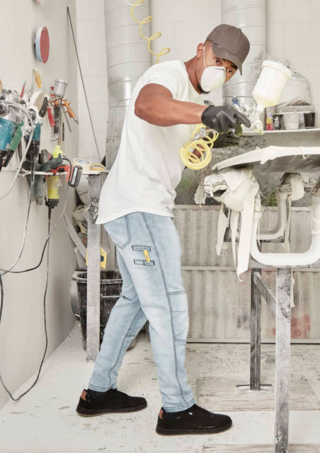A man wearing light blue stretch jeans and a white t-shirt is spray painting in a workshop.