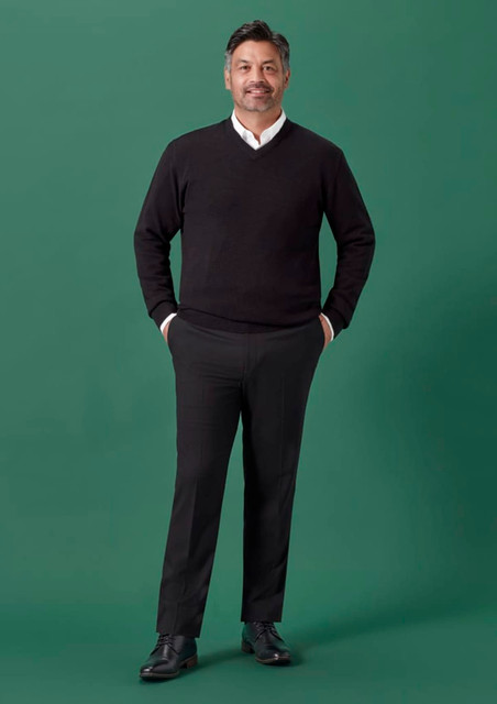 A man wearing a black sweater and black slim fit flat front pants stands against a green background.