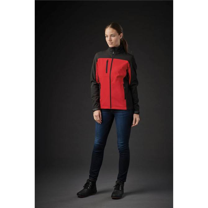 A woman is wearing a women's softshell jacket in red and black, with a zippered pocket, set against a dark background.