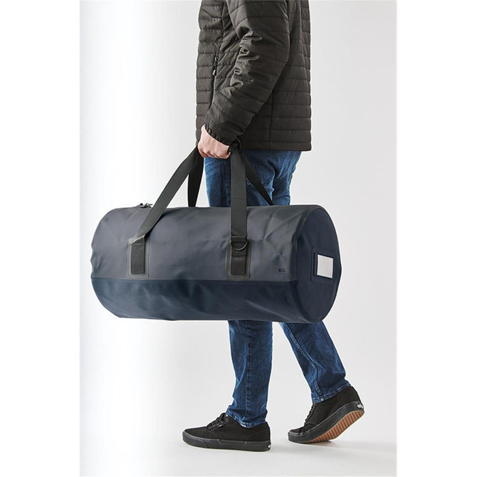 A person carrying a navy waterproof duffel bag with black straps and a branding logo.