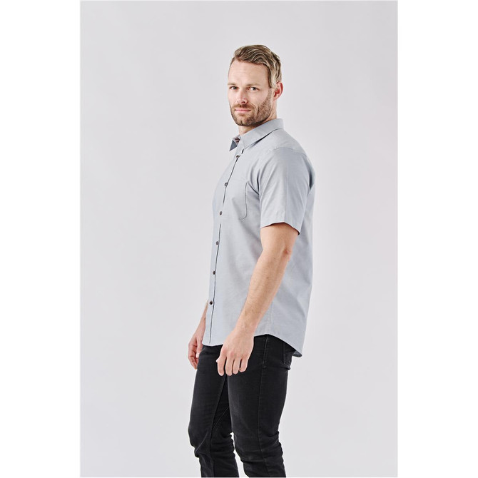 A light grey button-up shirt with short sleeves and a logo. The model is wearing black jeans.