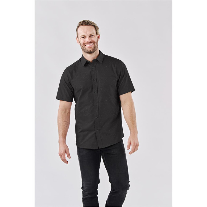 A man wearing a short-sleeved, black button-up shirt, standing against a light background. The shirt features a subtle pattern.