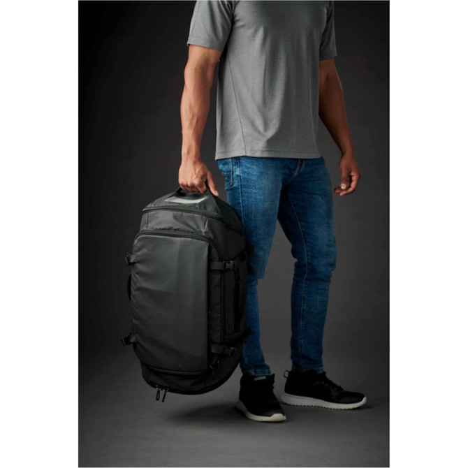 A man holding a dark green duffle bag, wearing a grey T-shirt and blue jeans, set against a dark background.