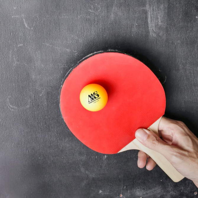 A table tennis paddle with a yellow ball resting on it, set against a dark background. The paddle features a logo.