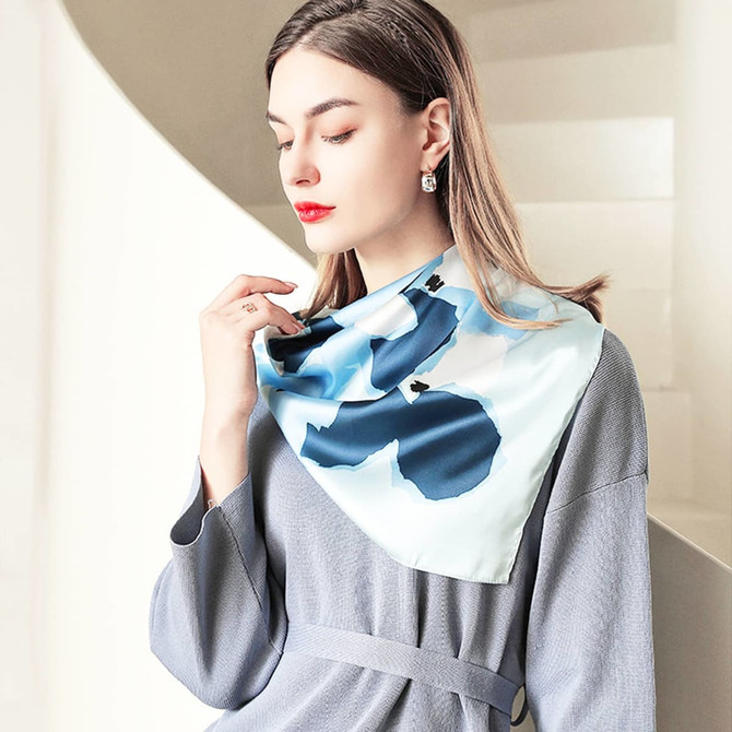 A silk chiffon scarf in light blue with abstract designs, displayed on a model wearing a grey outfit.